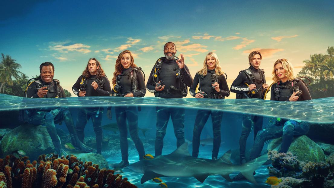 Seven celebrities in scuba gear, partially submerged in water with sharks below.