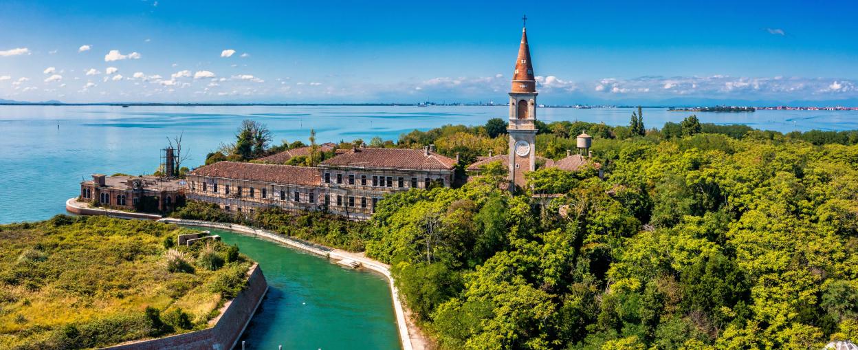 Aerial view of Poveglia Island in Venice, showing a dilapidated building and a clock tower surrounded by trees.