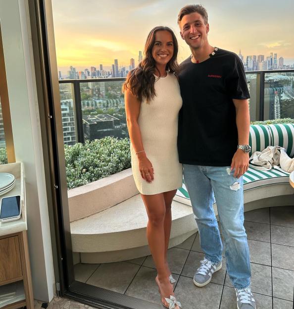 Desirae Krawczyk and Andrew Harris posing on a balcony overlooking a city skyline at sunset.