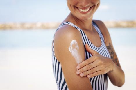 A cheerful woman with tan skin applies sun cream to her arm while wearing a striped swimsuit at a tropical Maldives beach under bright sunlight