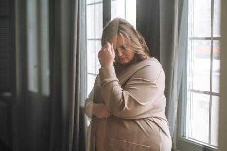 A plus-size woman stands by a window, looking sad.