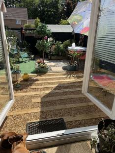 A dog sits by a patio door looking out at a sunny garden with gravel, wooden planks, and potted plants.