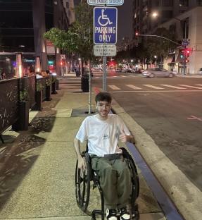 Man in wheelchair giving thumbs up in front of handicap parking sign.