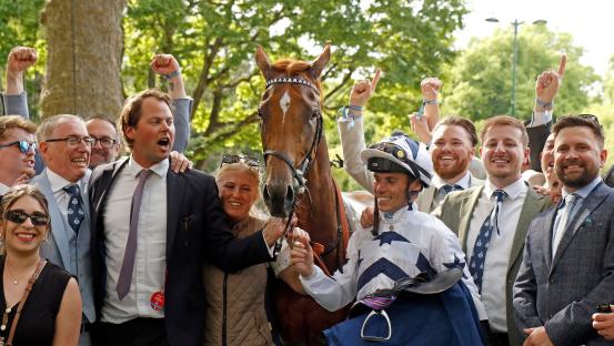 A horse and its winning jockey surrounded by jubilant owners and their team.
