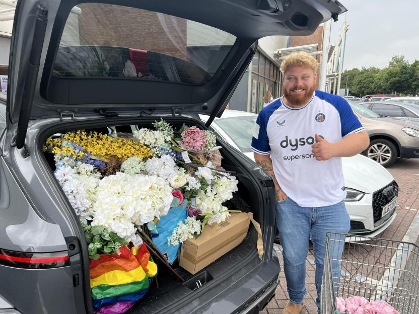 Man with thumbs up standing by his car trunk full of artificial flowers.