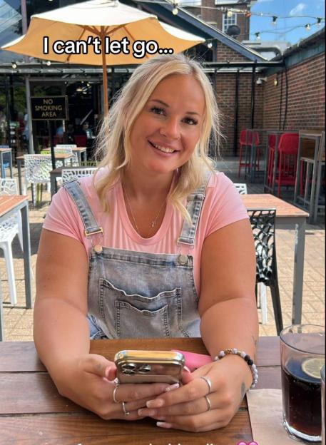 Woman in pink shirt and denim overalls sits at an outdoor table looking at her phone.
