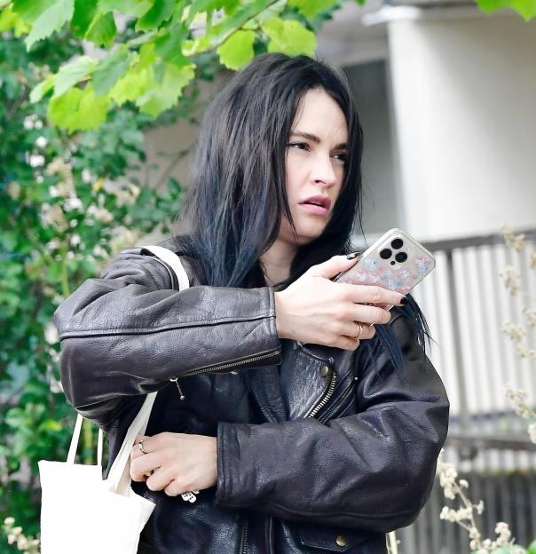 Lily James with dark hair, wearing a leather jacket, looks at her phone.