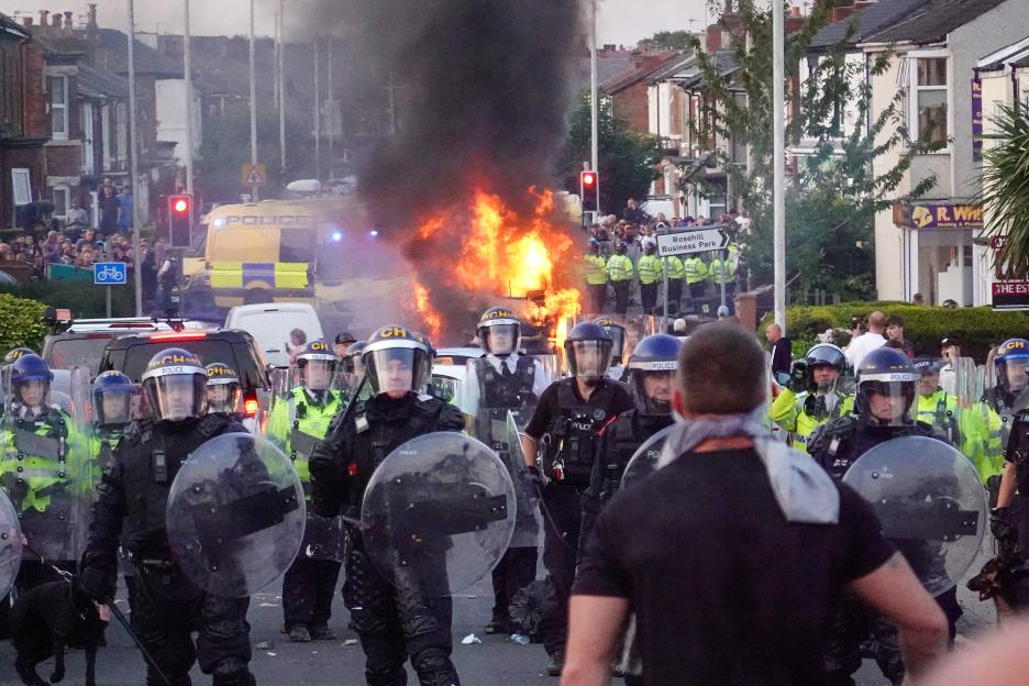 Riot police forming a line during a protest, a police vehicle on fire in the background.