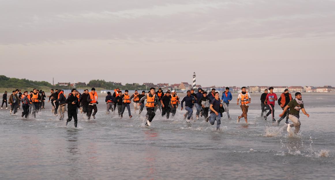 Migrants running across a beach toward a small boat.
