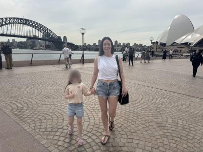 A woman and child walk hand-in-hand near the Sydney Opera House and Harbour Bridge.