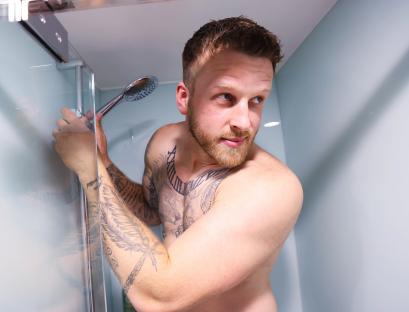A tattooed man adjusting a shower head in a small shower.