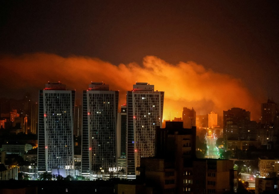 Nighttime view of city buildings with large orange smoke or fire in the background.