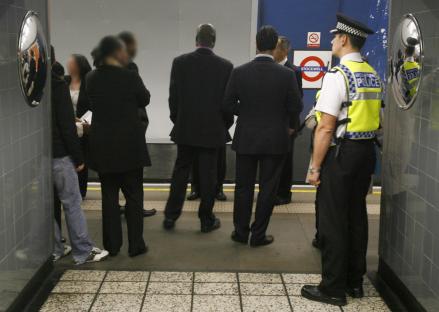 A police officer stands near a group of people whose faces are blurred, at a subway station.
