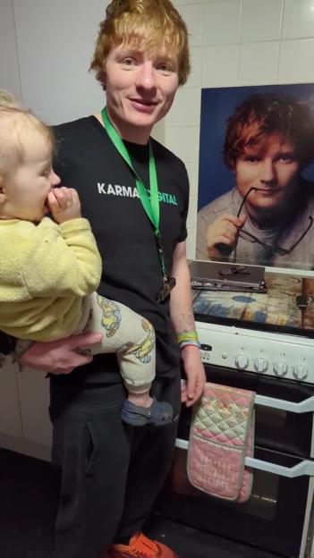 Man holding a baby, resembling Ed Sheeran, standing next to a poster of Ed Sheeran.