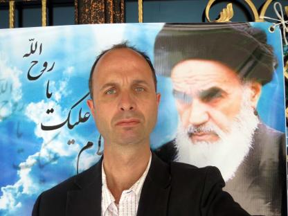 A man in a suit posing with a large photo of Ayatollah Khomeini in the background.