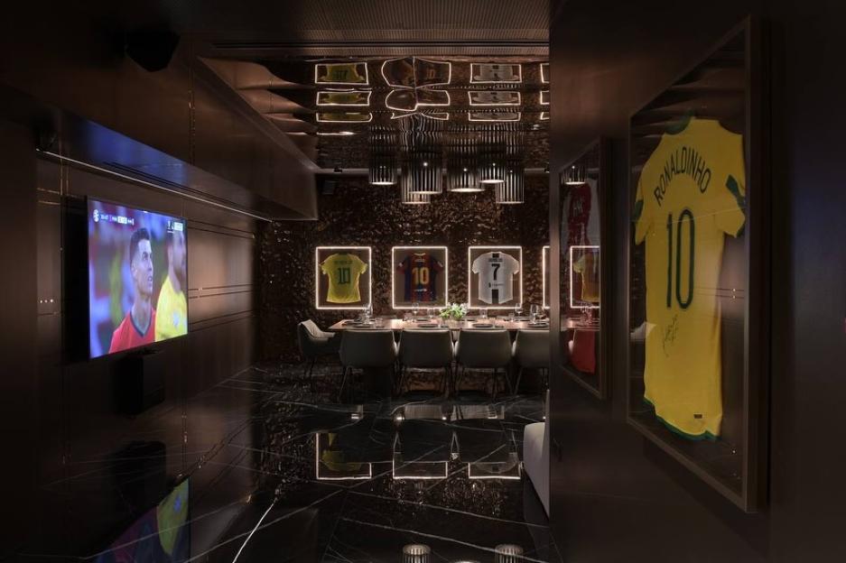 Interior view of a private dining room with framed soccer jerseys and a large screen showing a soccer game.
