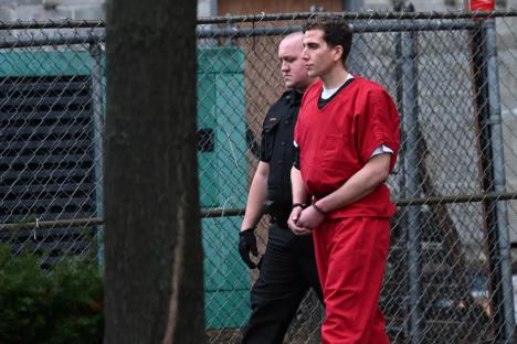 A handcuffed man in an orange jumpsuit is escorted by a guard.