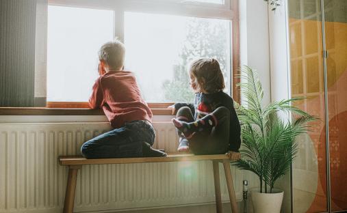 Two children sitting on a bench by a window, looking outside.