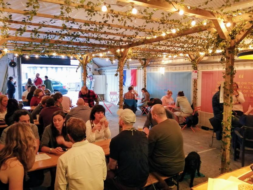 People seated at tables under a pergola decorated with ivy and string lights.