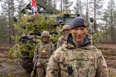 A British soldier in camouflage uniform, face camouflaged, stands in front of a military vehicle covered in branches.