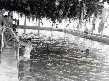 Black and white photo of people swimming in an indoor pool decorated with hanging plants.