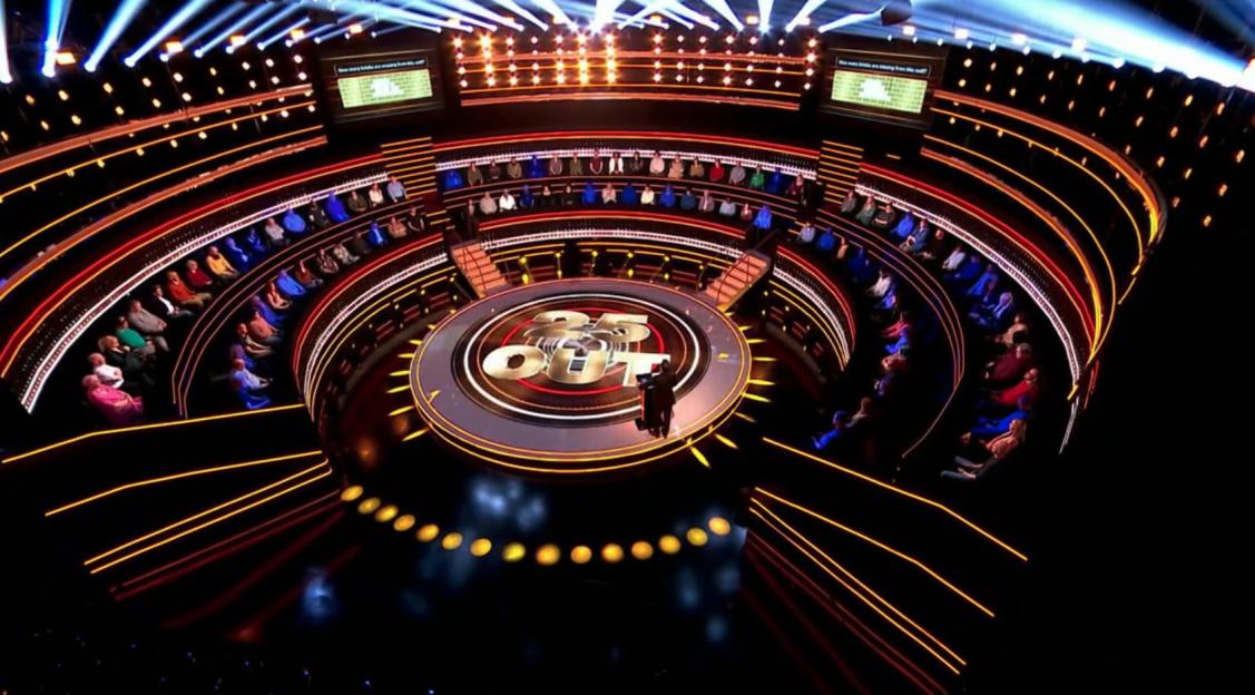 The 1% Club game show set with contestants seated in a circular tiered arrangement.