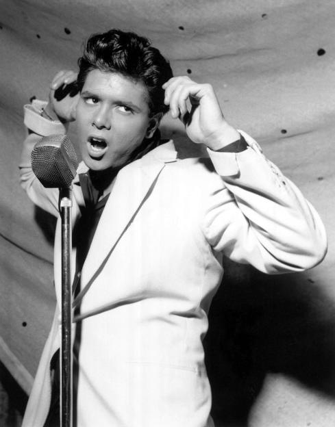Black and white photo of Cliff Richard singing into a microphone on stage.