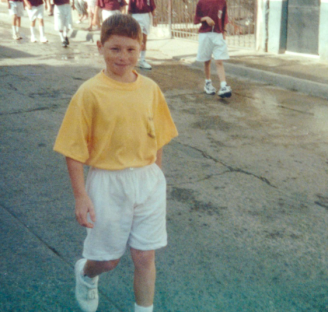 Young boy in yellow shirt and white shorts walking down a street.