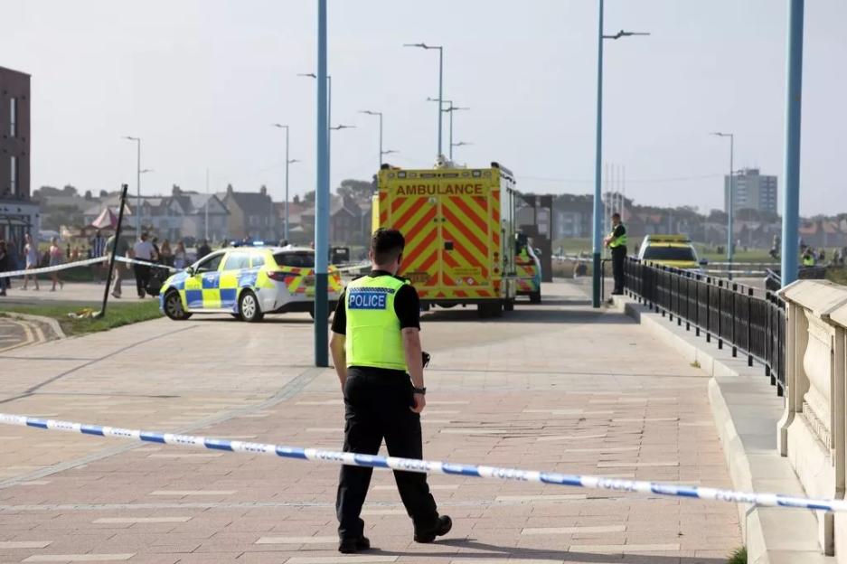 Police officer at a beach scene near an ambulance and police car, following a sea incident.