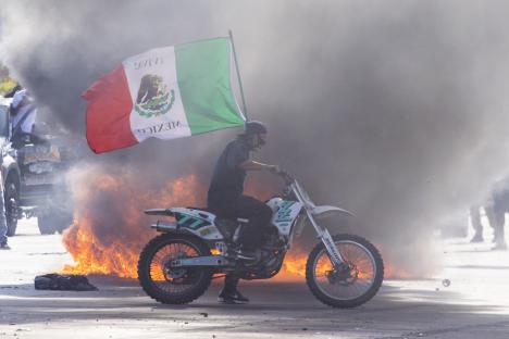 Person on a motorcycle with a Mexican flag during a protest with a burning car in the background.