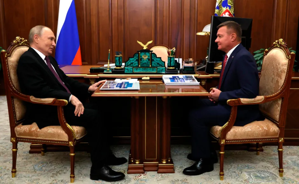 Vladimir Putin and another man in suits seated across a desk from each other.