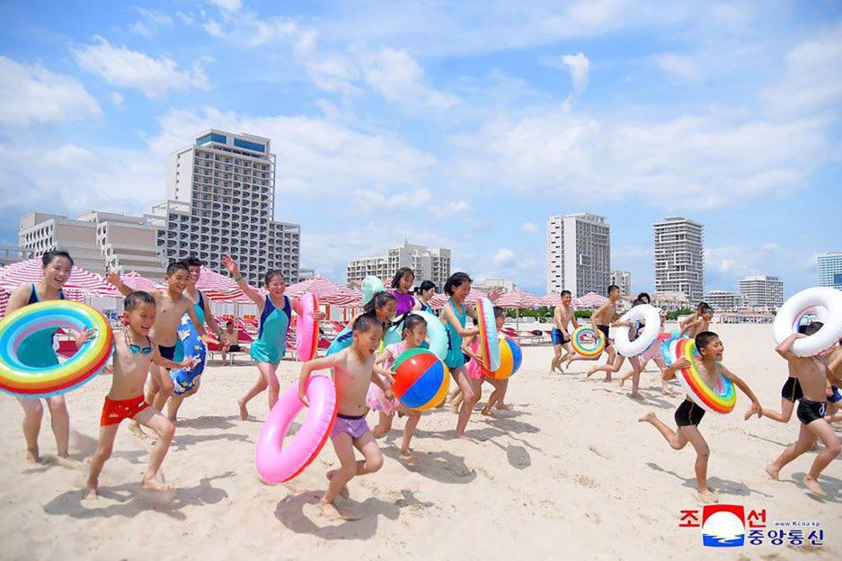 Children playing with inflatable rings and beach balls on a beach in front of hotels.