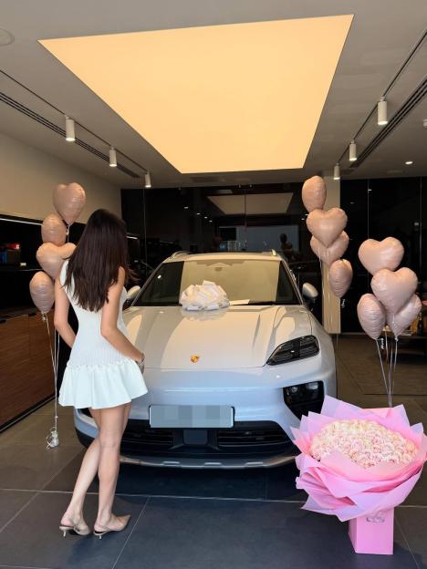 Woman in white dress stands next to a new Porsche with heart-shaped balloons and a large bouquet of roses.