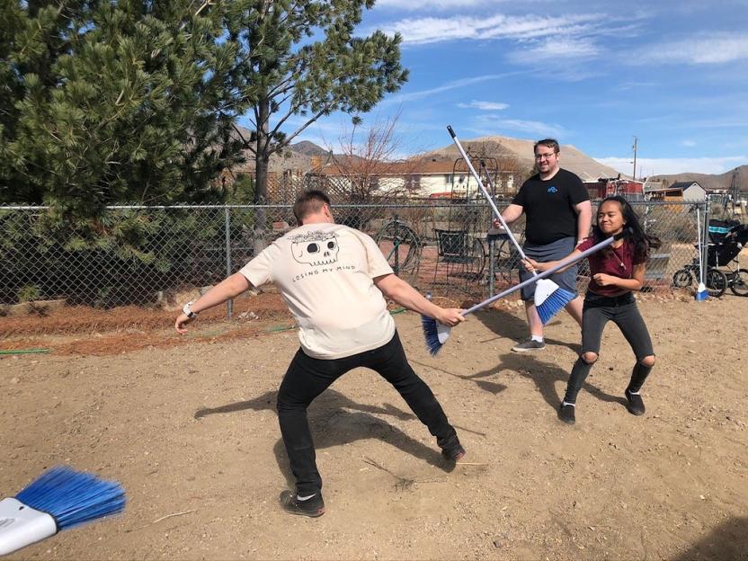Three people playfully duel with brooms in a yard.