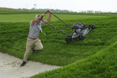 A groundskeeper struggles to pull a lawnmower up a steep grassy hill onto a sand trap.