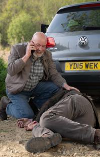 A man on a cell phone kneels beside an injured man lying on the ground.