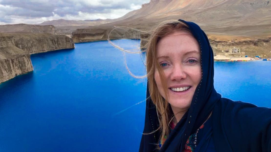 Woman in headscarf smiles at camera with blue lake and mountains in the background.