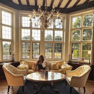 Woman sitting on a couch in a sunlit room with large windows and a chandelier.