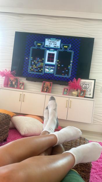 Legs of two people resting on a woven basket while watching a retro video game on a TV screen.