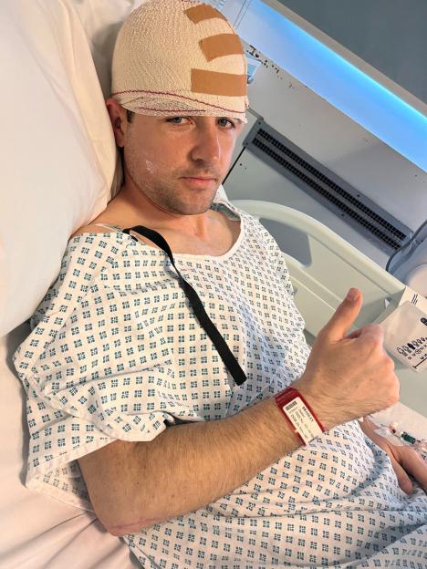 Man in hospital gown with head bandage giving thumbs up.