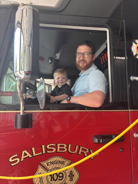 A man and a young boy sitting in the cab of a firetruck.