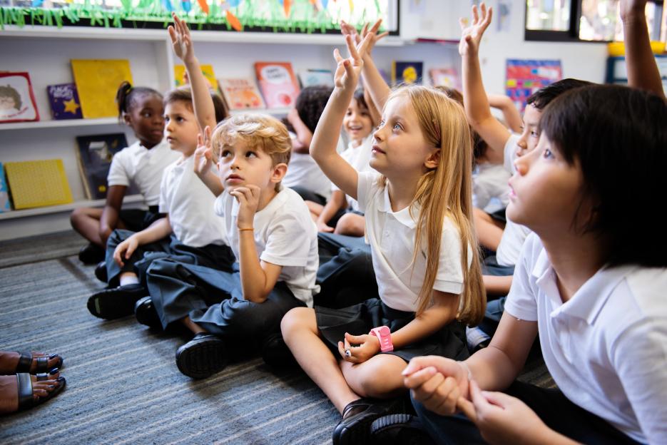 Diverse group of elementary school children sitting on the floor with their hands raised.