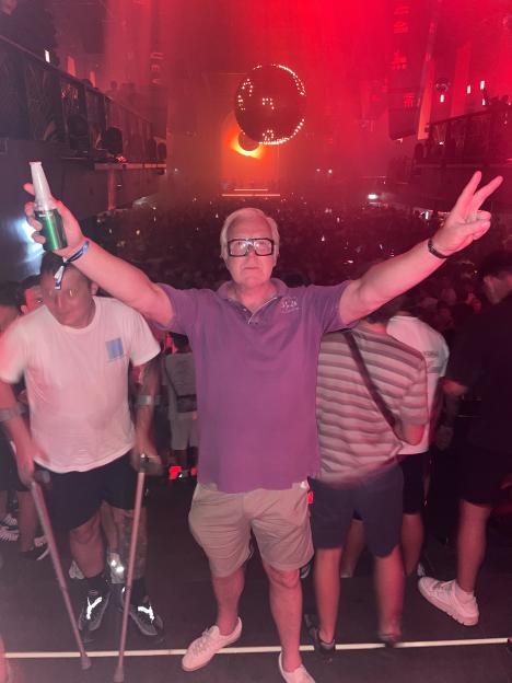 A man in a nightclub raises his arms, holding a beer bottle in one hand and giving a peace sign with the other.