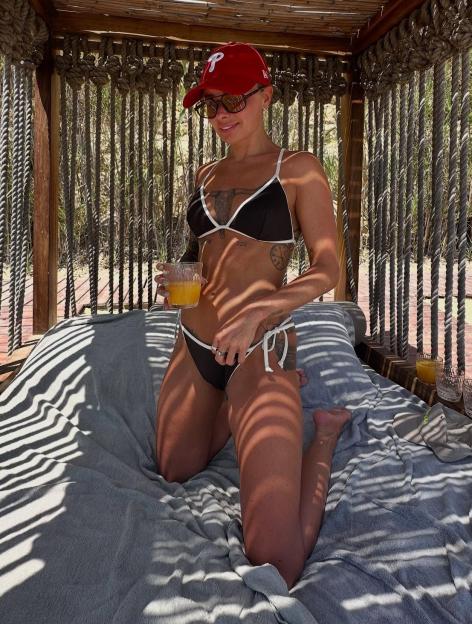 Woman in a bikini and baseball cap kneeling on a beach bed, holding a drink.