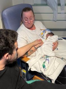A grandmother holds her premature granddaughter who is connected to medical equipment, while a man sits beside them.