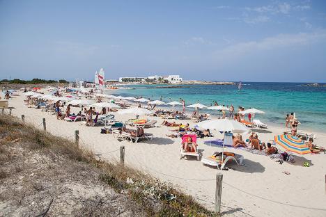 Crowded beach scene in Formentera with people sunbathing under umbrellas.