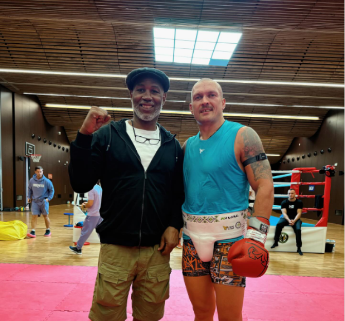 Two men in a boxing gym, one in boxing attire and the other in casual clothing.