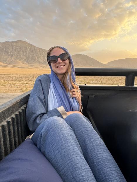 Woman in sunglasses and headscarf sitting in the back of a truck in a desert landscape.