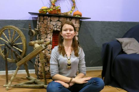 Woman sitting cross-legged in front of a faux fireplace and spinning wheel.