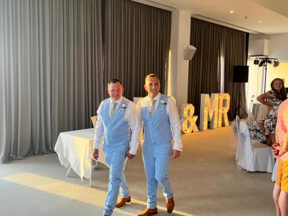 Two grooms in light blue suits walking down the aisle.
